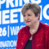 Woman in a red velvet jacket and pearl necklace smiling at a conference with a blue backdrop that reads 'Spring Meetings 2026'.