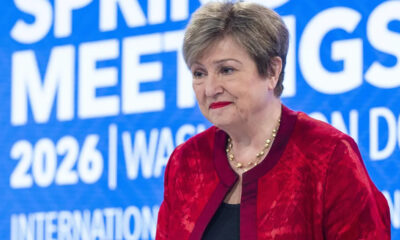Woman in a red velvet jacket and pearl necklace smiling at a conference with a blue backdrop that reads 'Spring Meetings 2026'.