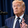 Former U.S. president Donald Trump speaking at a podium with a microphone, wearing a dark suit and tie.