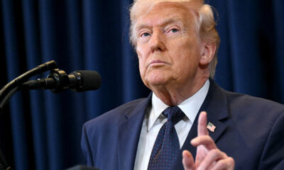 Former U.S. president Donald Trump speaking at a podium with a microphone, wearing a dark suit and tie.