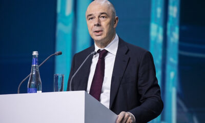 Man in a dark suit and burgundy tie speaking at a podium with microphones on stage