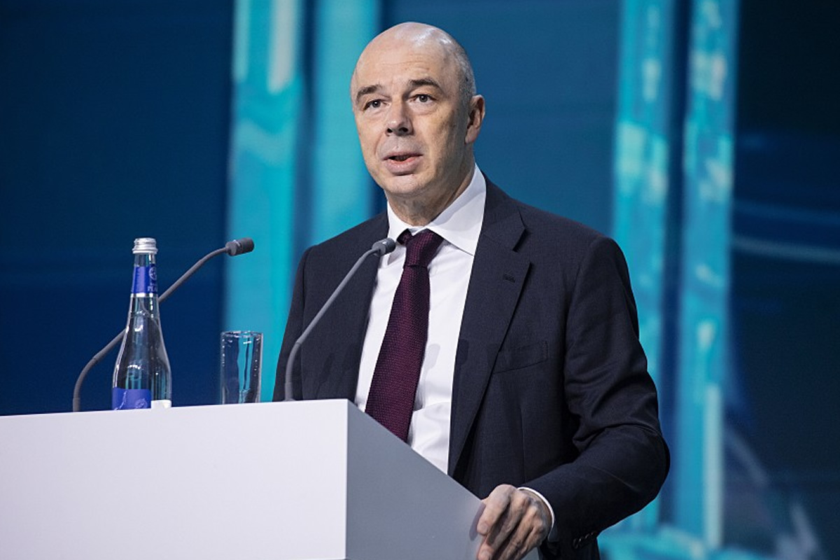 Man in a dark suit and burgundy tie speaking at a podium with microphones on stage