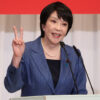 Asian woman in a blue blazer speaks at a podium, flashing a peace sign with her left hand, two microphones in front of her.