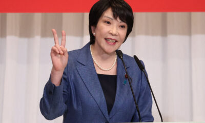 Asian woman in a blue blazer speaks at a podium, flashing a peace sign with her left hand, two microphones in front of her.