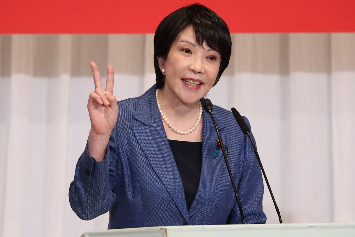 Asian woman in a blue blazer speaks at a podium, flashing a peace sign with her left hand, two microphones in front of her.