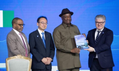 Four men in formal attire on a stage; one in a khaki uniform hands a booklet to another man in a suit during an awards ceremony.