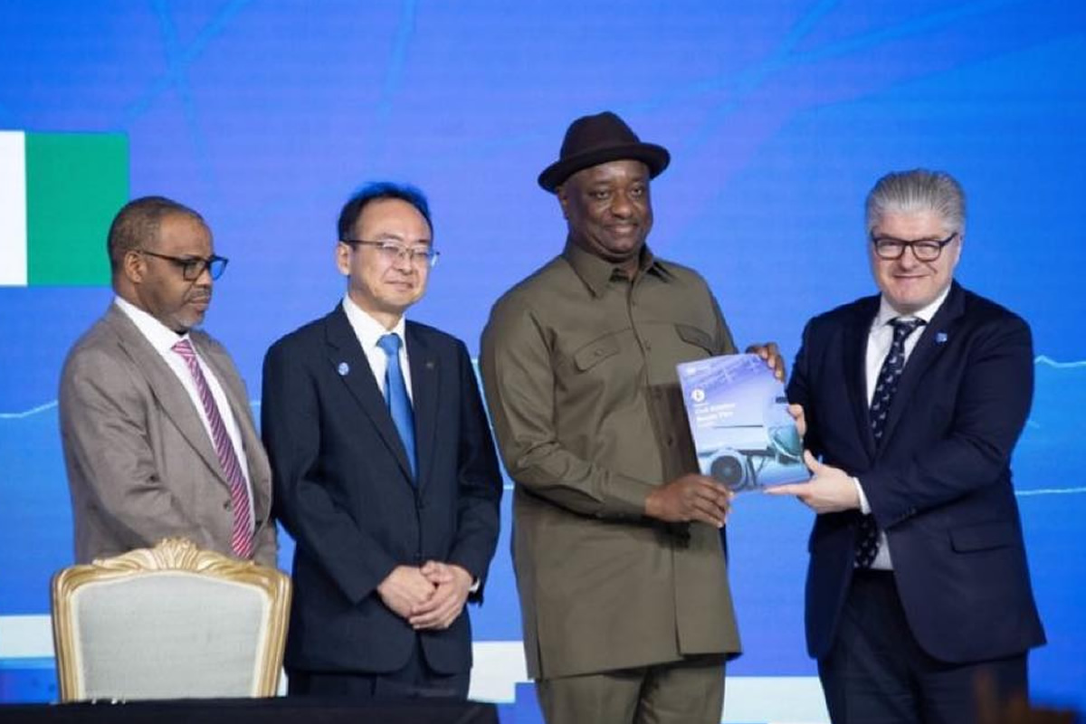 Four men in formal attire on a stage; one in a khaki uniform hands a booklet to another man in a suit during an awards ceremony.