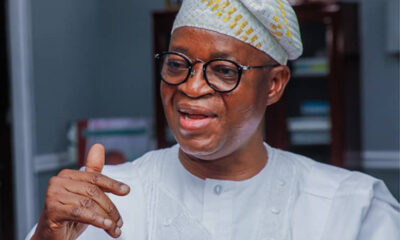 Close-up of a smiling man in a white traditional outfit and patterned cap, gesturing with his right hand as he speaks.