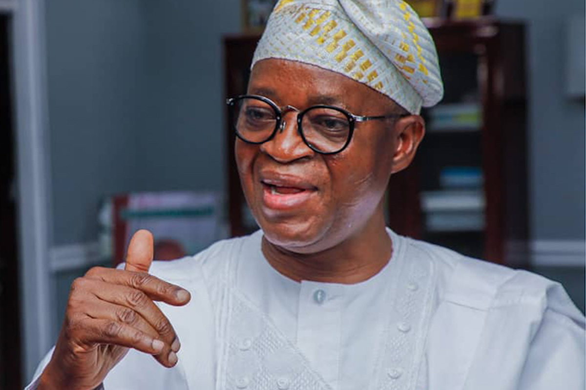 Close-up of a smiling man in a white traditional outfit and patterned cap, gesturing with his right hand as he speaks.